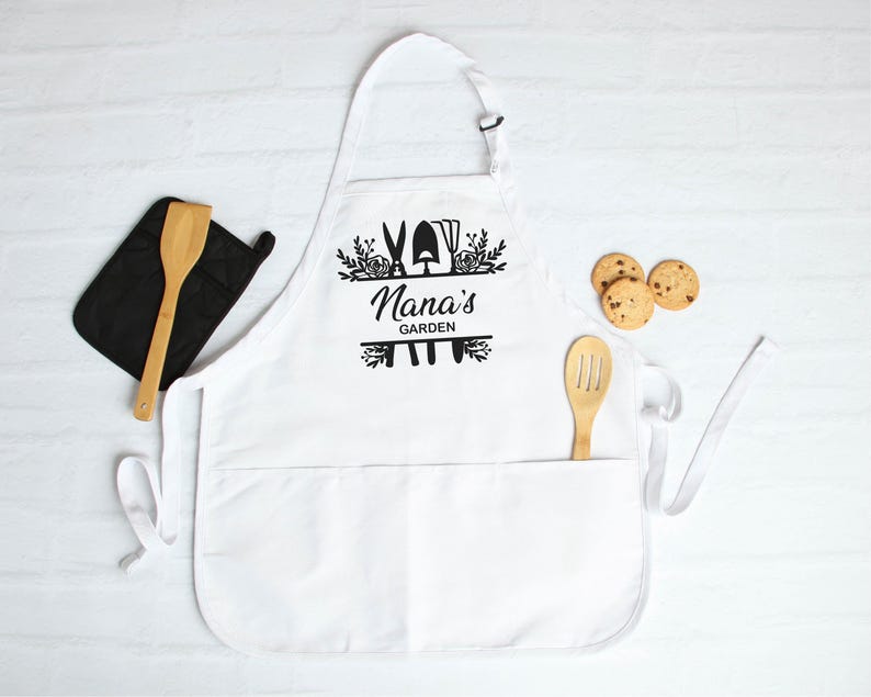 May include: White apron with a black design that says "Nana's Garden" with gardening tools and flowers. The apron has a large pocket in the front.
