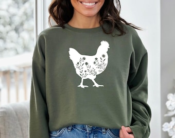 May include: An olive green crewneck sweatshirt featuring a large white graphic of a chicken silhouette. The interior of the chicken shape is filled with a detailed white line art illustration of various wildflowers, stems, and leaves.