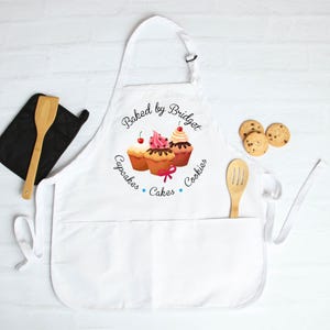 May include: White apron with a pocket, featuring a cupcake design and the text "Baked by Bridget". Includes a black oven mitt, wooden spoon, and chocolate chip cookies. A baking-themed gift.