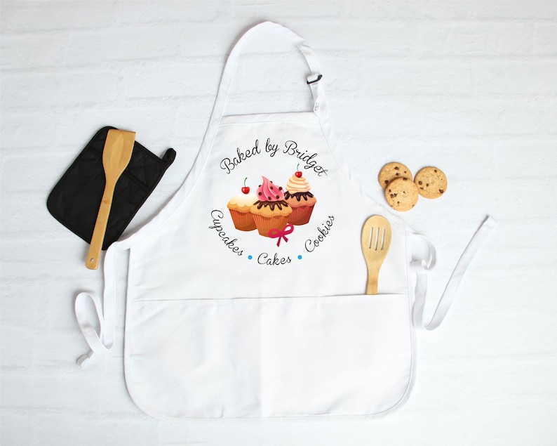 May include: White apron with a pocket, featuring a cupcake design and the text "Baked by Bridget". Includes a black oven mitt, wooden spoon, and chocolate chip cookies. A baking-themed gift.
