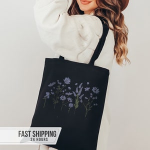 May include: Black tote bag featuring a floral design in shades of blue and green. The bag has a rectangular shape with two black handles. The floral design includes various flowers and foliage. The bag is made of a sturdy fabric.