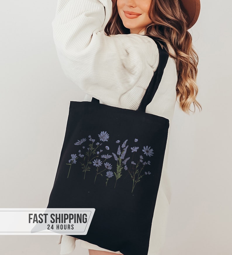May include: Black tote bag featuring a floral design in shades of blue and green. The bag has a rectangular shape with two black handles. The floral design includes various flowers and foliage. The bag is made of a sturdy fabric.