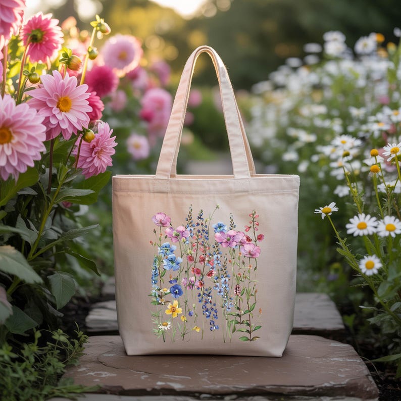 A beige tote bag with a floral design printed on it, placed on a stone path surrounded by a variety of colorful flowers.