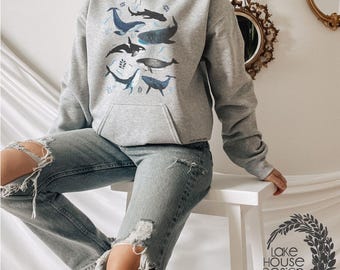 May include: A light gray hooded sweatshirt featuring a graphic print of various whales and sharks in shades of blue, black, and white. The hoodie includes a front kangaroo pocket, long sleeves, and a drawstring hood, paired with distressed blue jeans.