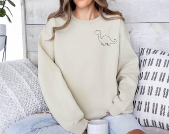 May include: A beige crewneck sweatshirt featuring a small, simple black line art illustration of a dinosaur on the upper left chest area. The garment has long sleeves with ribbed cuffs and a ribbed neckline, providing a clean and straightforward look.