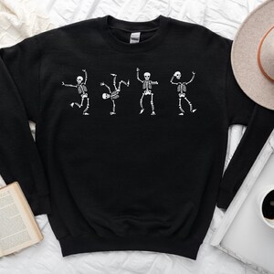 Puede incluir: Sudadera negra con un gr&aacute;fico blanco de cuatro esqueletos bailando. Los esqueletos est&aacute;n en varias poses, con los brazos y las piernas extendidos. La sudadera est&aacute; hecha de un material suave.