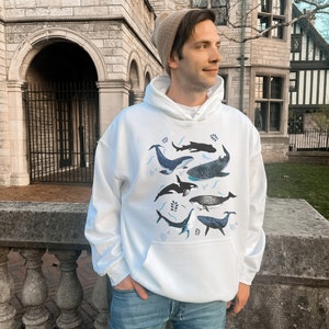 May include: White hoodie featuring a graphic print of different whales and dolphins in shades of blue and grey. The hoodie has a drawstring hood and a kangaroo pocket.