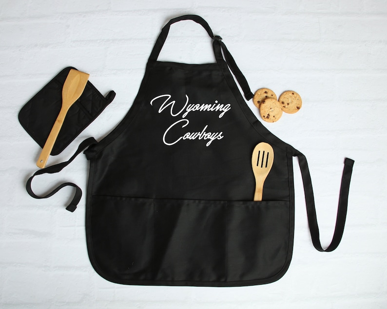 May include: Black kitchen apron with two pockets and a white embroidered design that says "Wyoming Cowboys". The apron is shown with a wooden spoon and a pot holder.