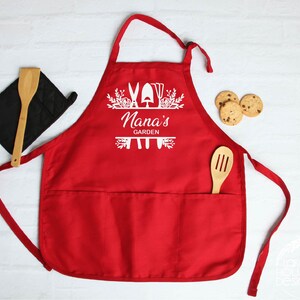 May include: Red gardening apron with a white design that says "Nana's Garden" with a floral border and gardening tools.