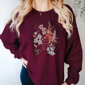 Pu&ograve; includere: Felpa color borgogna con stampa floreale con fiori blu, rosa e rossi. I fiori sono disposti in un design simile a un bouquet.