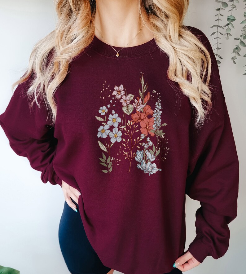 Pu&ograve; includere: Felpa color borgogna con stampa floreale con fiori blu, rosa e rossi. I fiori sono disposti in un design simile a un bouquet.