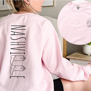 May include: Pink sweatshirt with the word "NASHVILLE" printed vertically down the back, with a pair of black cowboy boots on either side of the word. The front of the sweatshirt is plain pink.
