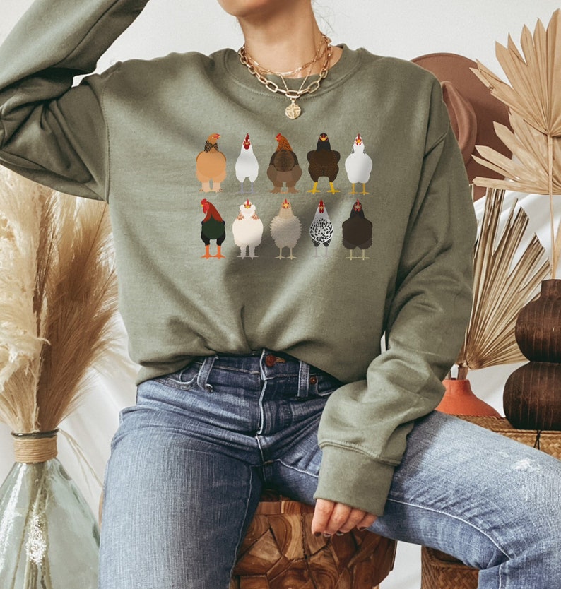 May include: A green sweatshirt with a crew neck and a graphic of 10 chickens in different colors and patterns. The sweatshirt is being worn by a person wearing blue jeans.