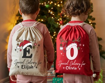 May include: Two drawstring Christmas gift bags, one beige and one red, with festive designs. The beige bag features a reindeer antler and Santa hat graphic with the letter "E" and the text "Special Delivery for Elijah". The red bag has a similar design with the letter "O" and the text "Special Delivery for Olivia".