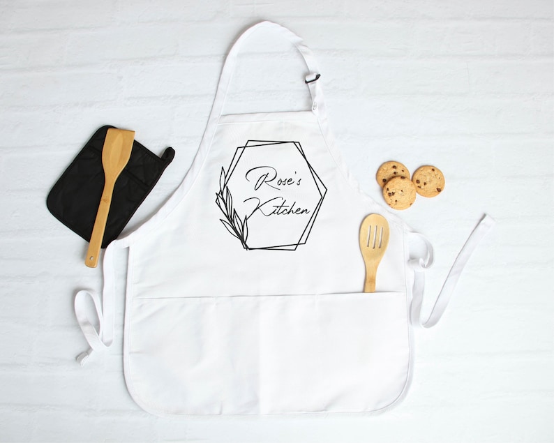 May include: White kitchen apron with a black geometric design and the text "Rose's Kitchen".