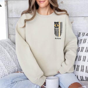 May include: A light beige sweatshirt with a black and yellow graphic of a flag with the text "DISGRACIFIER" and the name "Kelsey" printed below.