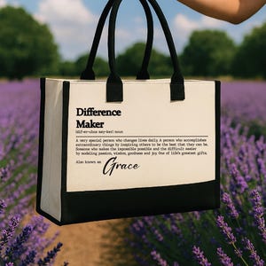 May include: A canvas tote bag with black handles and a black bottom panel. The bag features the text "Difference Maker" and a definition, with "Grace" written below. The background shows a field of purple flowers.