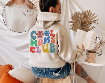 May include: A beige hooded sweatshirt featuring a large, colorful graphic on the back with the text "COOL MOMS CLUB" in a wavy, retro-style font, accented by flower icons. A smaller matching graphic is printed on the front left chest area.