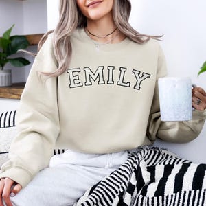 May include: A beige crewneck sweatshirt featuring the name EMILY printed in a black collegiate-style font across the chest. The garment has long sleeves and a ribbed neckline, cuffs, and hem, providing a simple and clean aesthetic.