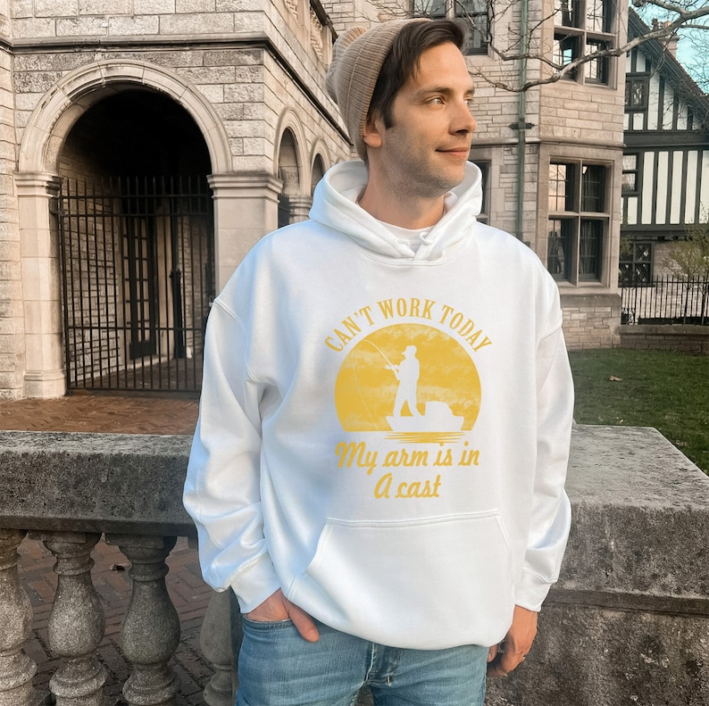 May include: White hooded sweatshirt with a yellow graphic of a person fishing in a boat. The graphic says "Can't work today, my arm is in a cast."