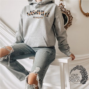 May include: A gray hooded sweatshirt with the text "Nashville Tennessee" in a yellow and brown patterned font.