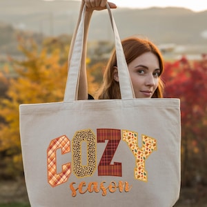 A woman holding a large tote bag with the word "COZY" printed on it, standing in front of a scenic autumn landscape.
