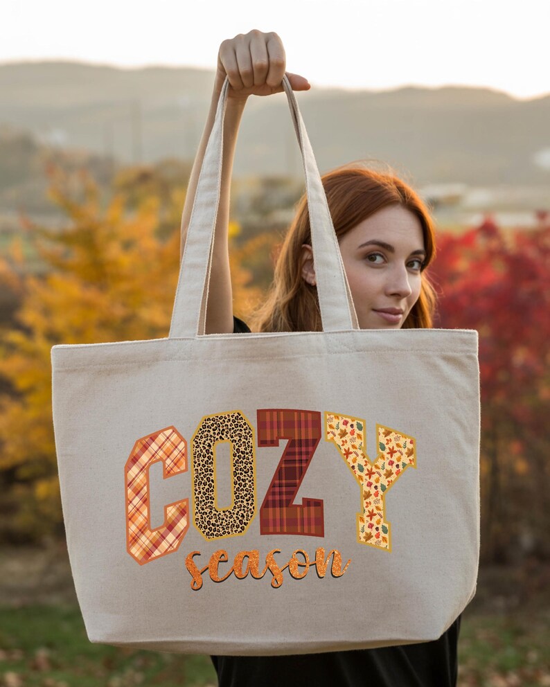 A woman holding a large tote bag with the word "COZY" printed on it, standing in front of a scenic autumn landscape.
