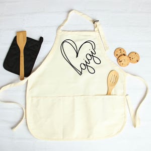May include: A white cotton apron with a large black heart and the word "Gigi" written inside. The apron has a large pocket on the front.