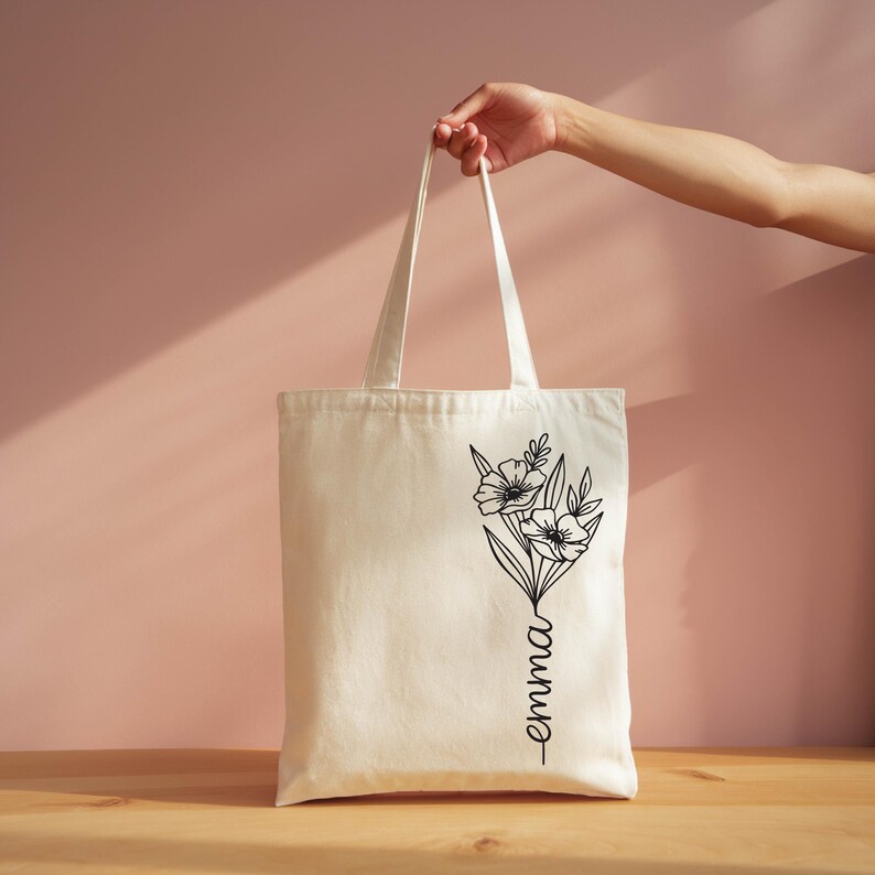 A hand holding a white tote bag with a floral design on it, against a pink background.