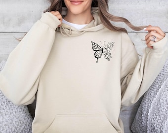 May include: A beige hooded sweatshirt featuring a black line art graphic on the left chest. The design depicts a butterfly where one wing is patterned with traditional butterfly markings and the other wing is composed of various floral elements.