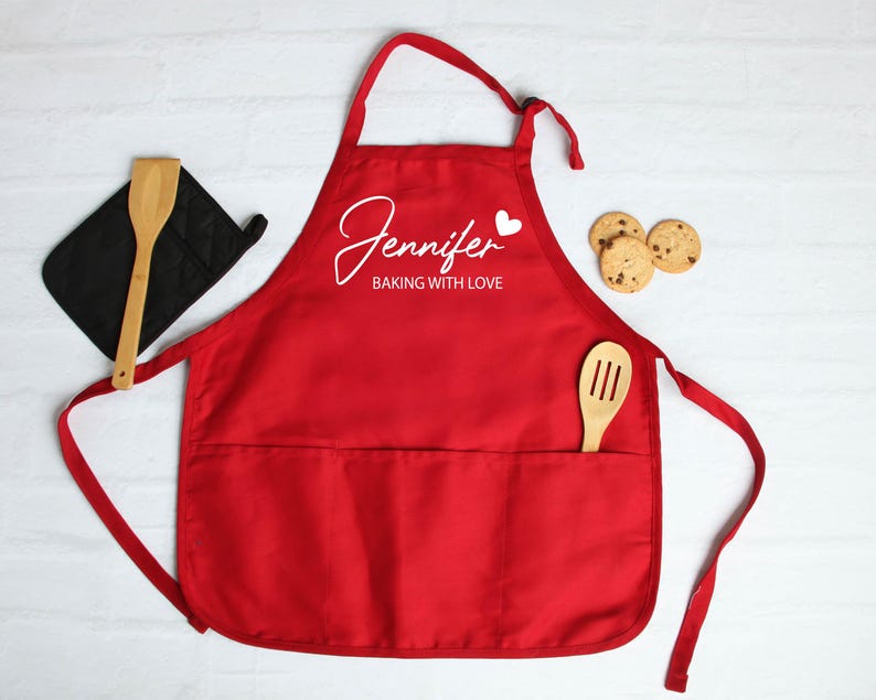 Peut inclure: Un tablier rouge avec le nom "Jennifer" et la phrase "Baking with Love" en &eacute;criture blanche. Une cuill&egrave;re en bois et une manique noire sont &agrave; gauche. Une spatule en bois est dans la poche, et trois biscuits sont &agrave; droite.
