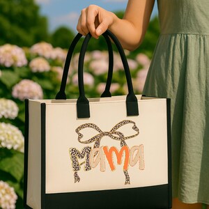 A person is holding a large tote bag with the word "Mama" printed on it, standing in front of a field of white flowers.