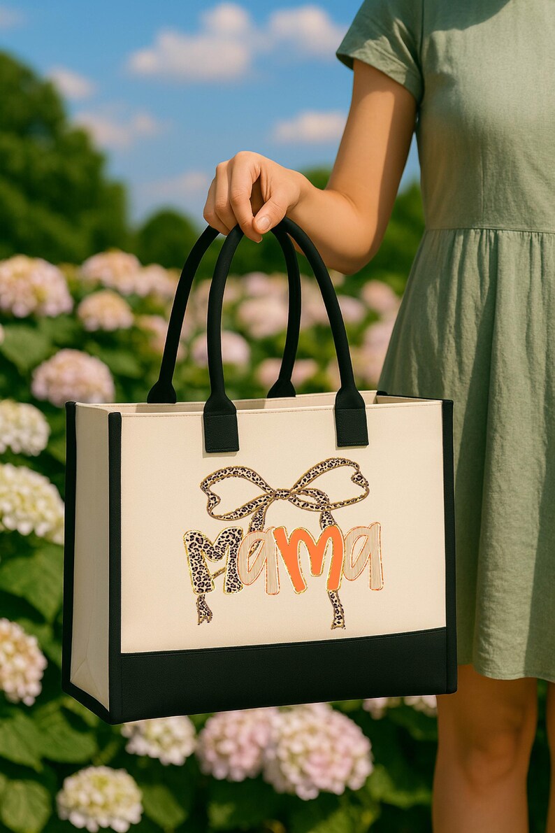 A person is holding a large tote bag with the word "Mama" printed on it, standing in front of a field of white flowers.