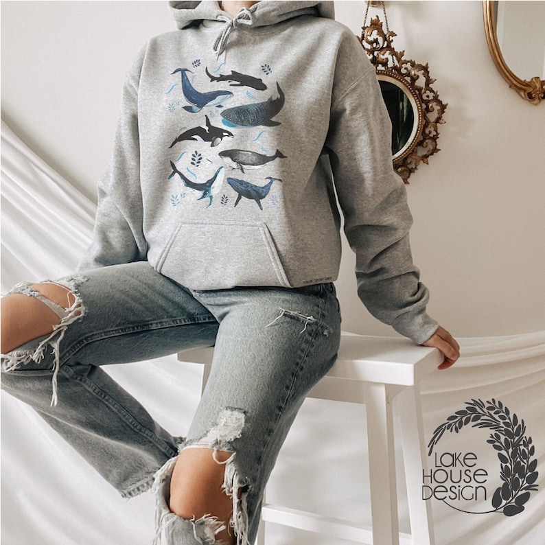 May include: A grey hoodie featuring a graphic print of different whales and dolphins in shades of blue. The hoodie has a drawstring hood and a pouch pocket.