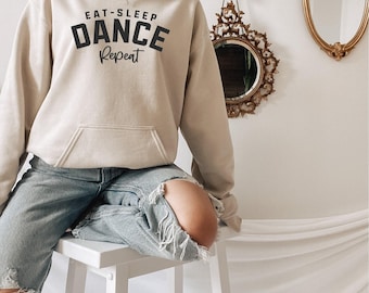 May include: A beige hooded sweatshirt featuring the black text EAT-SLEEP DANCE Repeat printed on the chest. The garment includes a front kangaroo pocket, long sleeves, and a drawstring hood. The hoodie is paired with light blue distressed denim jeans.