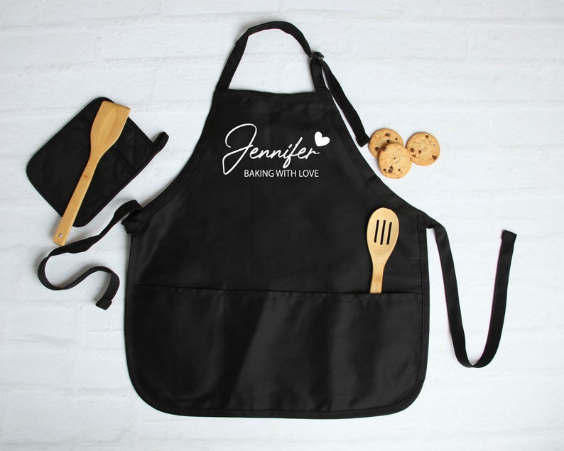 Peut inclure: Tablier noir avec le nom "Jennifer" et la phrase "Baking with Love" en &eacute;criture blanche. Une cuill&egrave;re en bois et une manique sont &agrave; c&ocirc;t&eacute; du tablier. Des cookies aux p&eacute;pites de chocolat sont &eacute;galement dans l'image. Le tablier a deux poches.