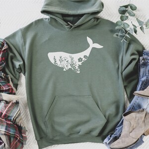 May include: A green hooded sweatshirt with a white whale silhouette filled with floral designs. The sweatshirt is laid out on a white surface with a plaid scarf and blue jeans with brown boots.
