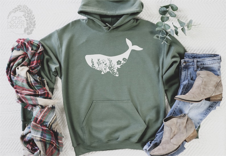May include: A green hooded sweatshirt with a white whale silhouette filled with floral designs. The sweatshirt is laid out on a white surface with a plaid scarf and blue jeans with brown boots.