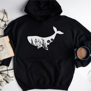 May include: Black hoodie with a white whale silhouette filled with floral designs. The hoodie has a drawstring hood and a kangaroo pocket.