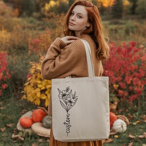 A woman with red hair stands in a field of autumn foliage, holding a white tote bag with a floral design.