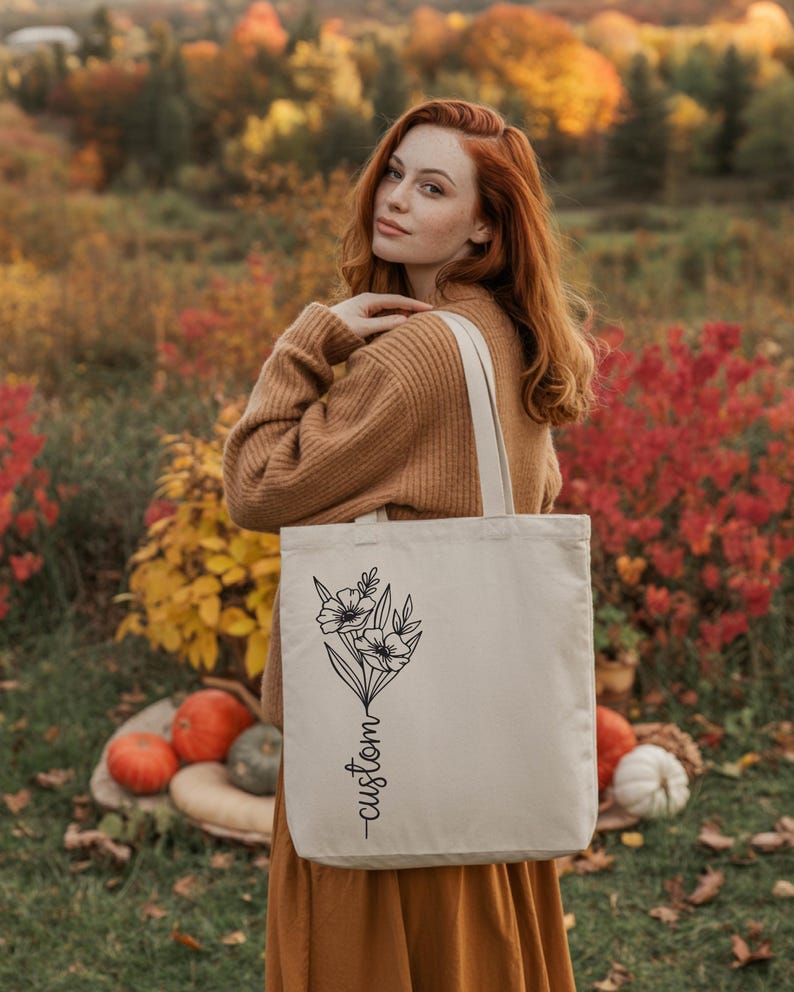 A woman with red hair stands in a field of autumn foliage, holding a white tote bag with a floral design.