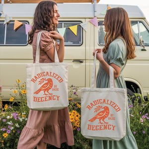 May include: Two women holding off-white tote bags with the text "RADICALIZED BY BASIC DECENCY" in orange. The bags feature an orange bird graphic with an olive branch. The women are standing in front of a van decorated with colorful flags.