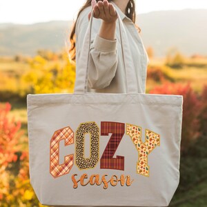 A woman holding a large tote bag with the word "COZY" printed on it, standing in a field with autumn foliage in the background.
