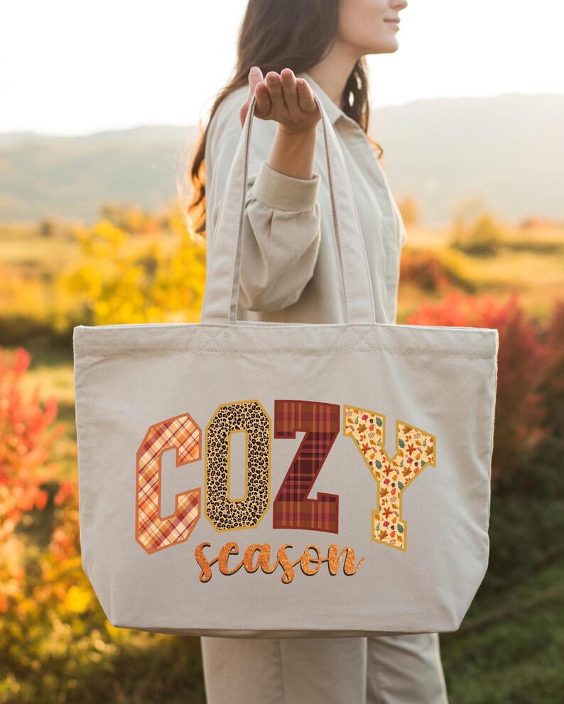 A woman holding a large tote bag with the word "COZY" printed on it, standing in a field with autumn foliage in the background.