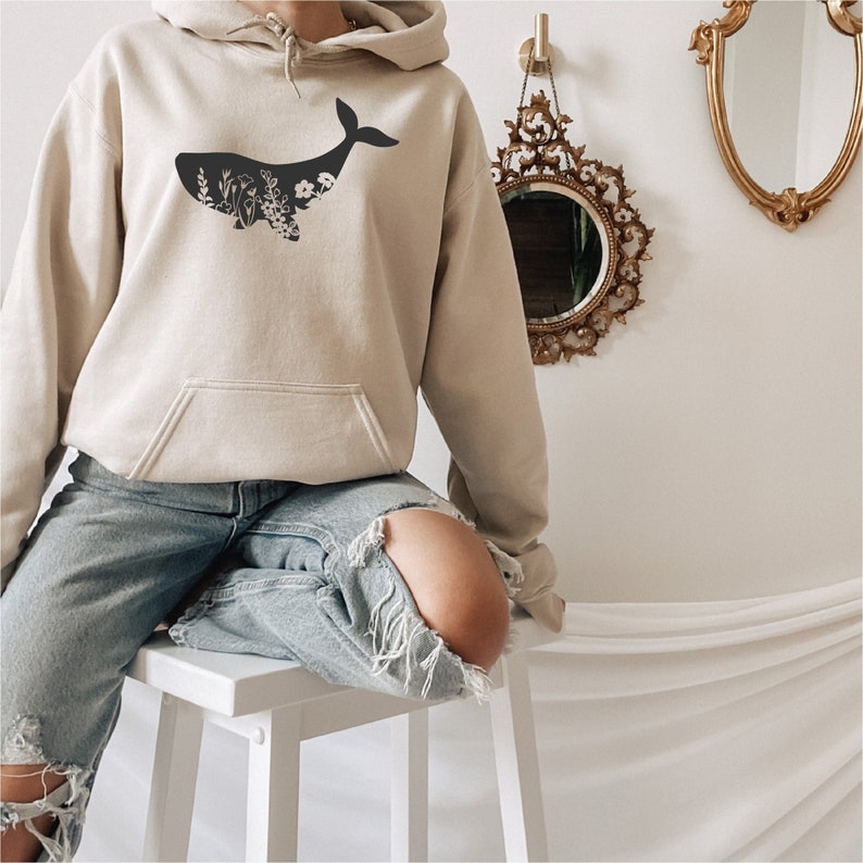 May include: A beige hoodie with a black silhouette of a whale with floral details inside the whale's body. The model is wearing the hoodie and blue jeans.