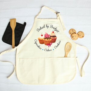 May include: Cream-colored apron with a pocket, featuring a cupcake design and the text "Baked by Bridget, Cupcakes, Cakes, Cookies." A wooden spoon rests in the pocket. A black oven mitt and cookies are also present.