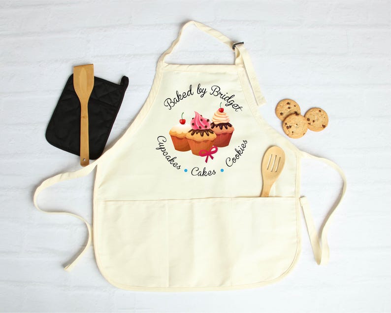 May include: Cream-colored apron with a pocket, featuring a cupcake design and the text "Baked by Bridget, Cupcakes, Cakes, Cookies." A wooden spoon rests in the pocket. A black oven mitt and cookies are also present.