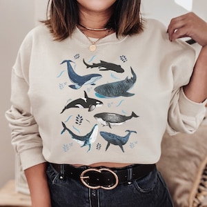 May include: A beige cropped sweatshirt featuring a graphic print of different whales and ocean-inspired designs. The sweatshirt has a relaxed fit and a crew neckline.