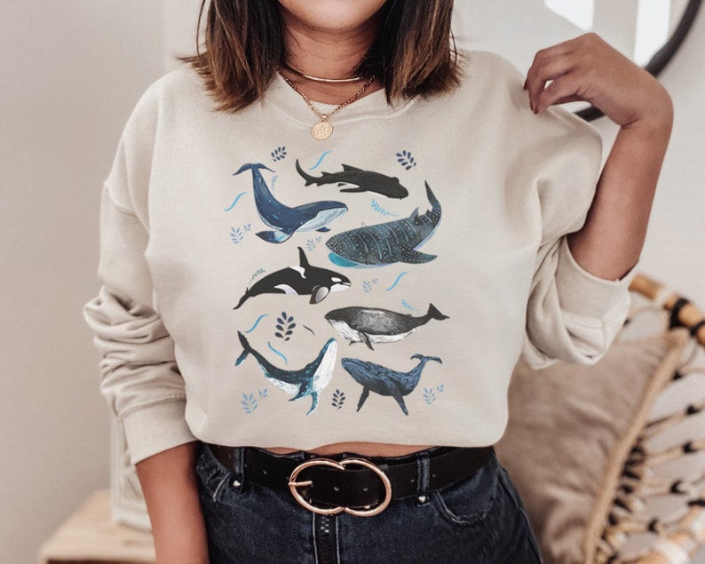 May include: A beige cropped sweatshirt featuring a graphic print of different whales and ocean-inspired designs. The sweatshirt has a relaxed fit and a crew neckline.