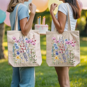 Two women standing in a grassy area, each carrying a large tote bag with a floral pattern. In the background, there is a cake with candles, suggesting a celebration or special occasion.